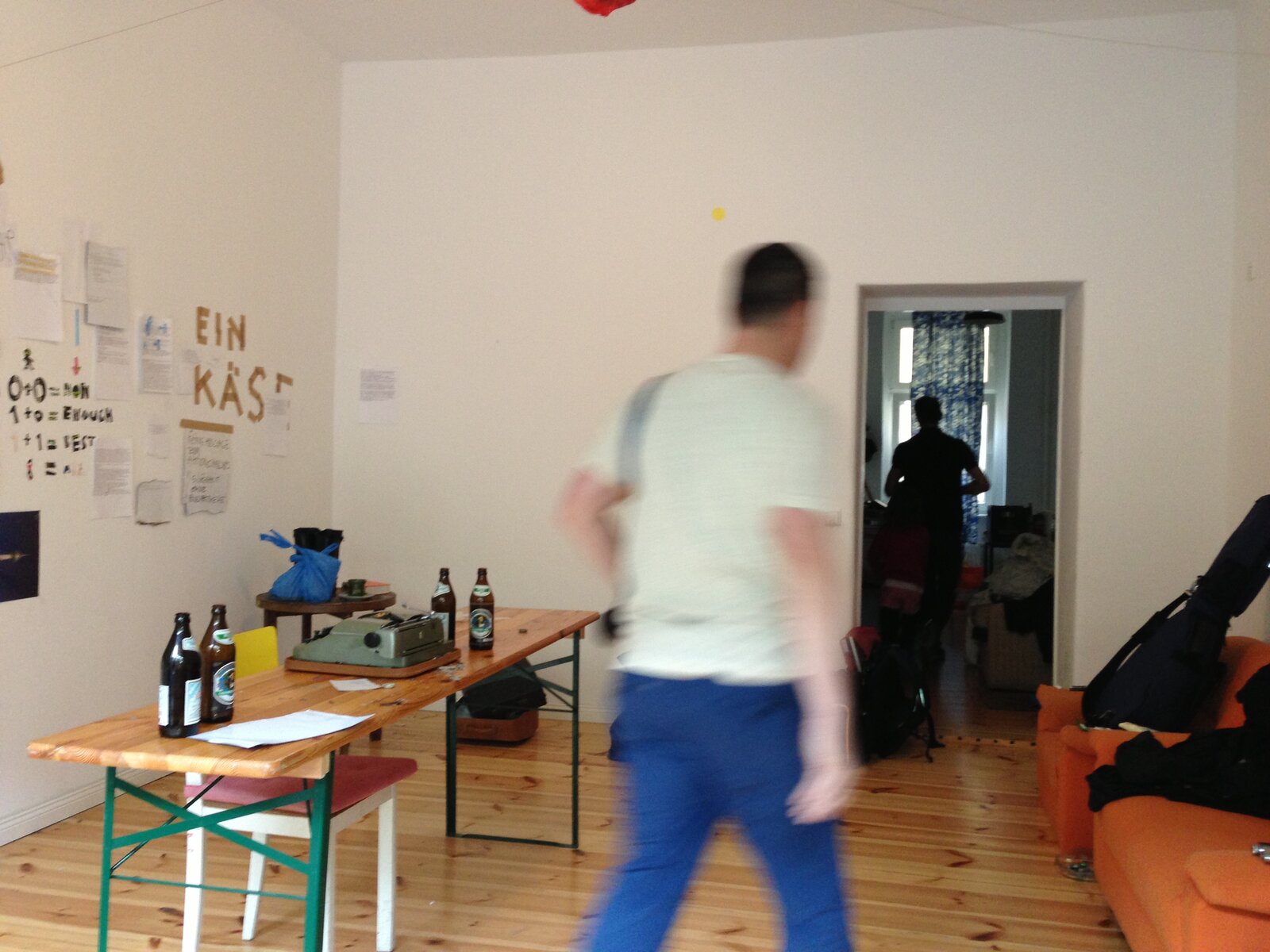 A room with a formula on the wall, '0+0=non 1+0=enough 1+1=best', a typewriter and beer bottles on a table, someone walking away in motion blur