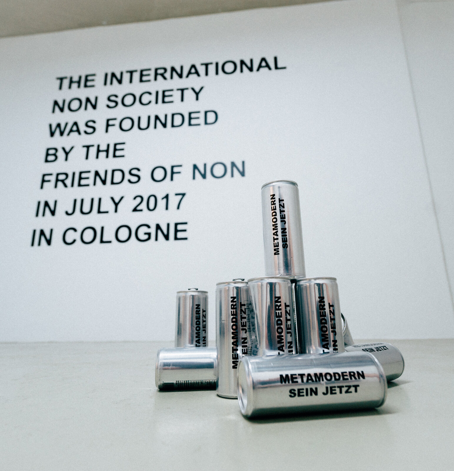 Aluminium cans reading 'METAMODERN SEIN JETZT' in front of a wall text: 'The International NON Society was founded by the Friends of NON in July 2017 in Cologne'