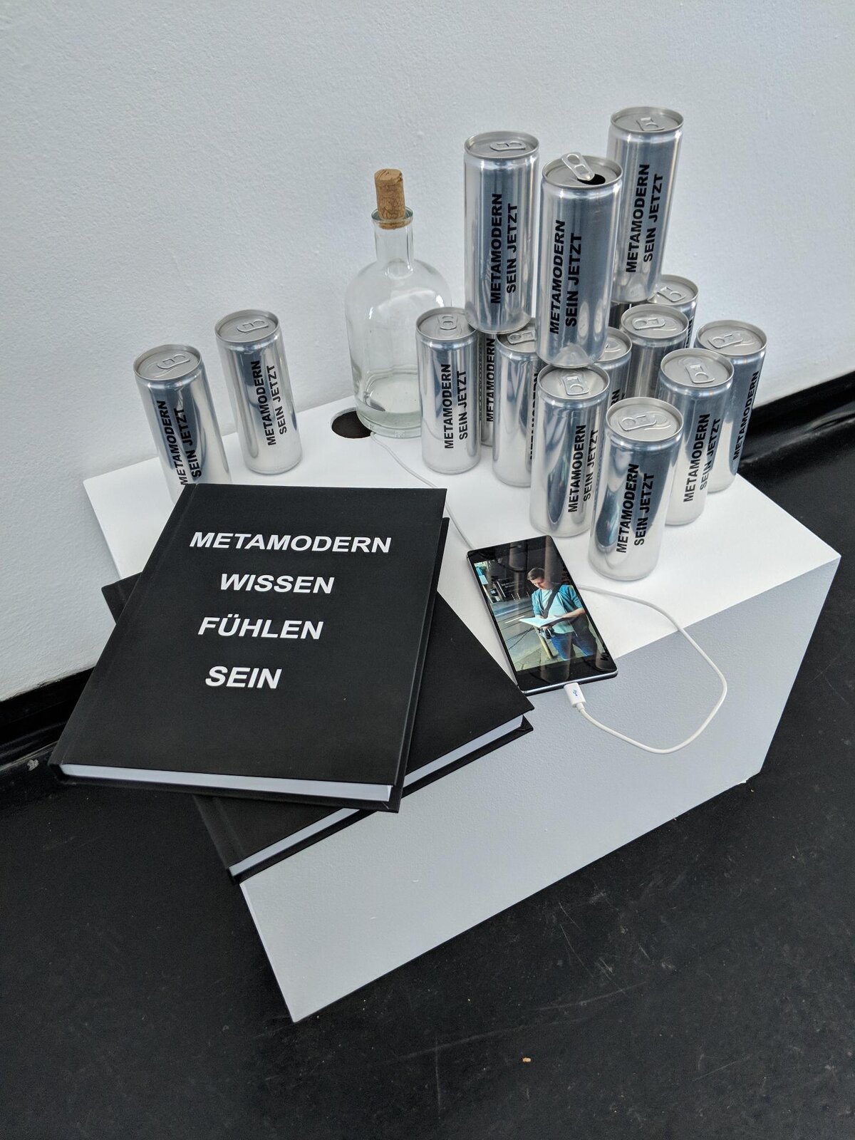 Installation view: 'METAMODERN SEIN JETZT' cans and copies of METAMODERN WISSEN FÜHLEN SEIN on a plinth, a phone playing a photo propped against the books