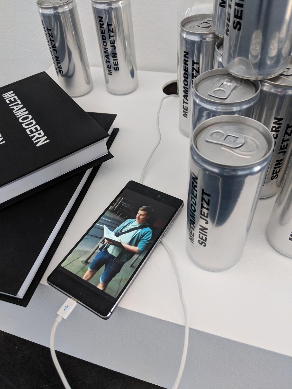 Closer view of the same plinth: cans, books and a phone showing a photo of Florian Kuhlmann reading