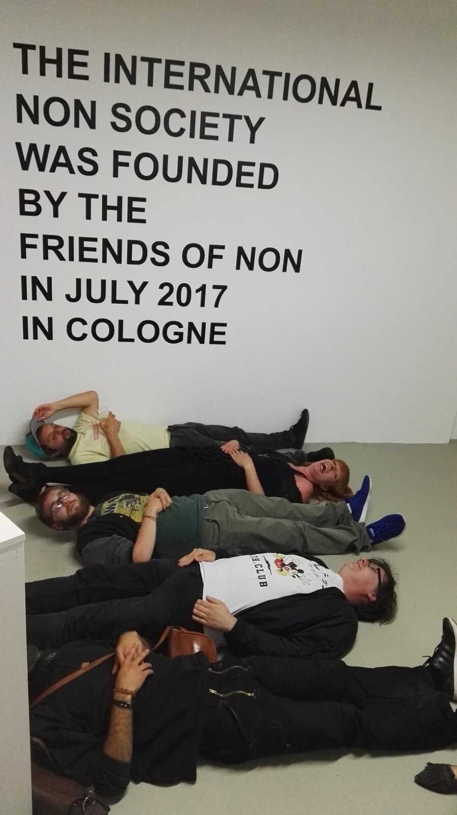 A wall reading 'THE INTERNATIONAL NON SOCIETY WAS FOUNDED BY THE FRIENDS OF NON IN JULY 2017 IN COLOGNE', people lying on the floor beneath it, laughing
