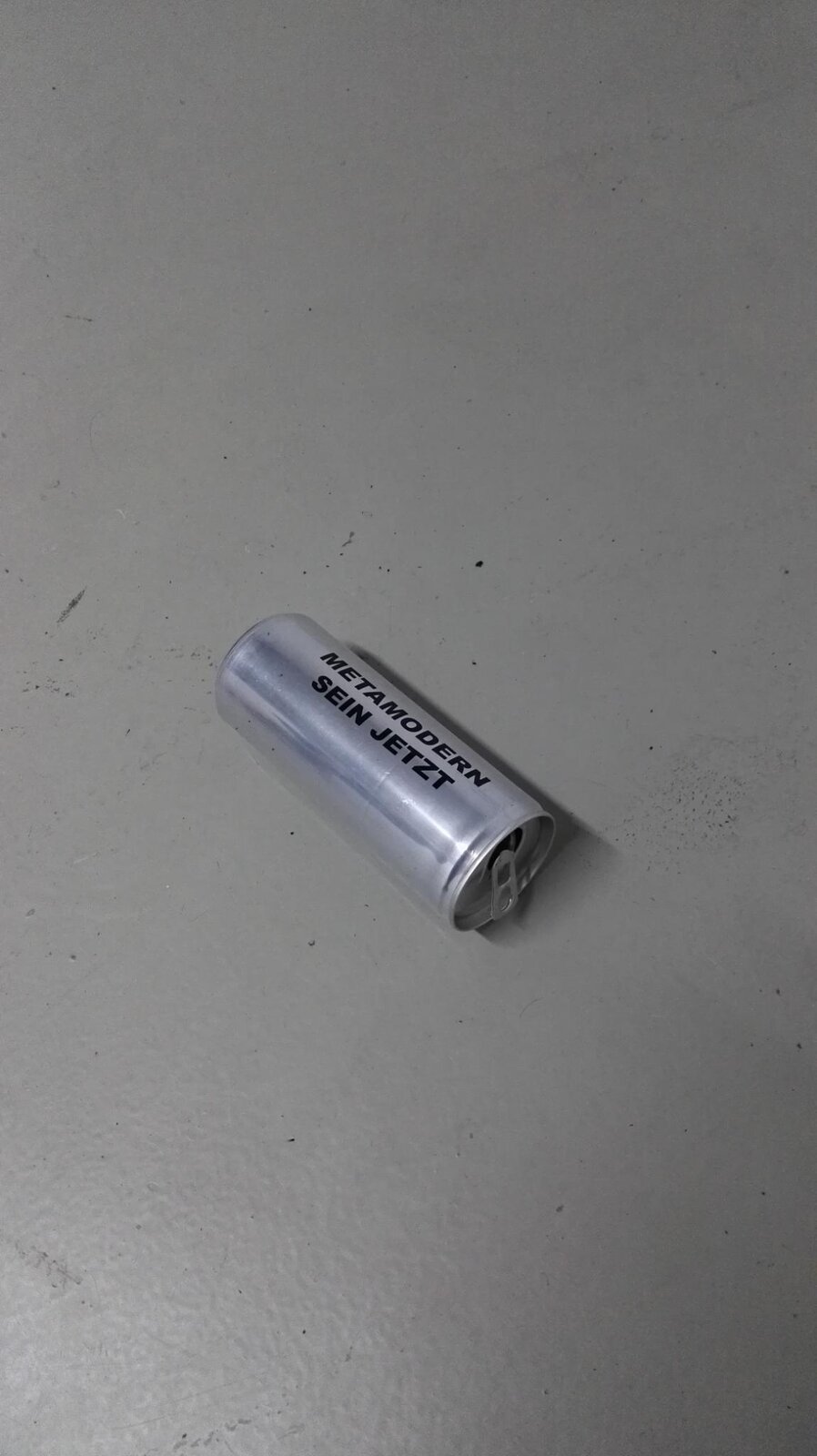 A single 'METAMODERN SEIN JETZT' can lying on a bare concrete garage floor