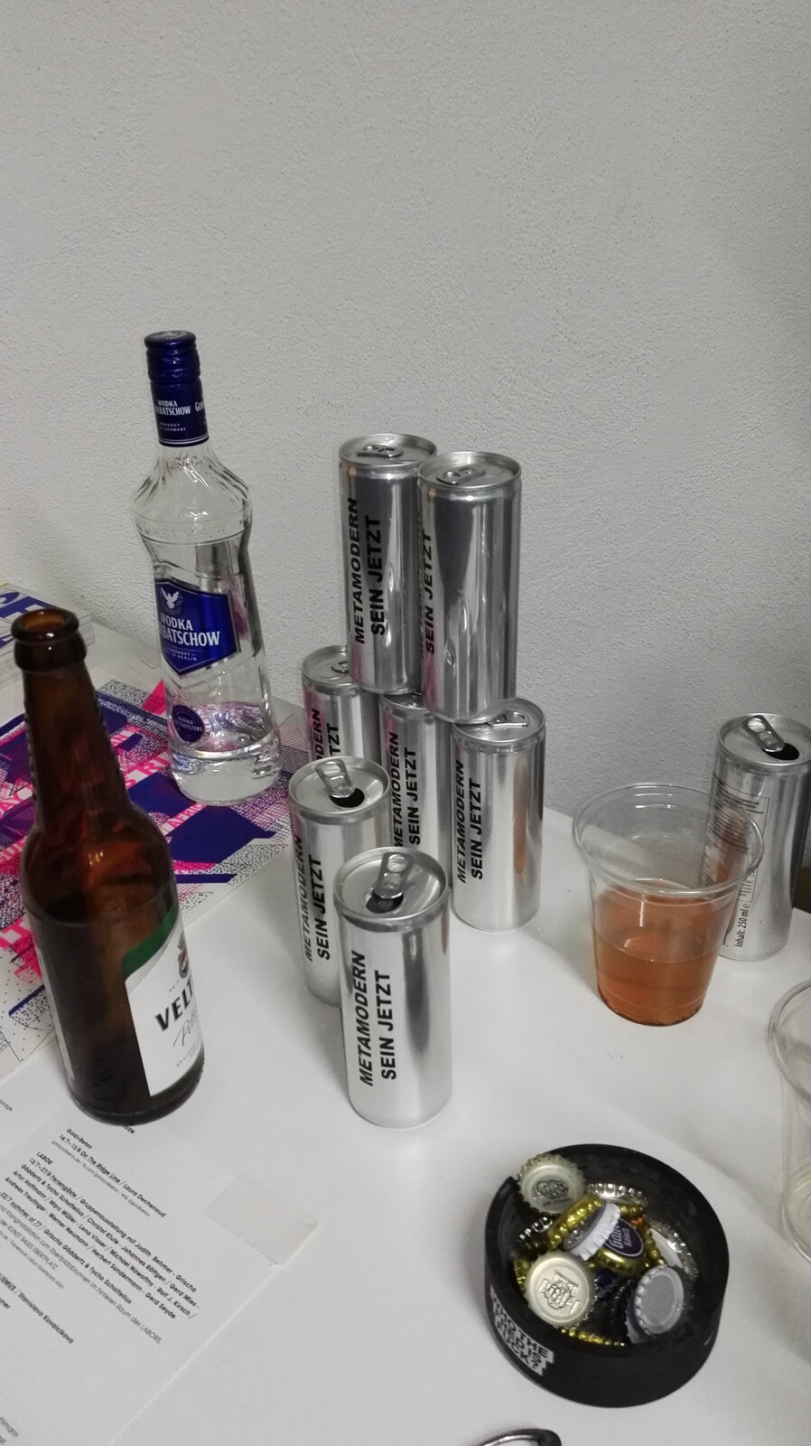 A row of aluminium cans reading 'METAMODERN SEIN JETZT' on a table next to a vodka bottle, a Veltins beer bottle and a plastic cup of orange-coloured mix