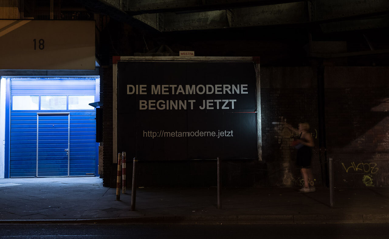 A black poster under a railway bridge at night reading 'DIE METAMODERNE BEGINNT JETZT — http://metamoderne.jetzt', a passerby walking by
