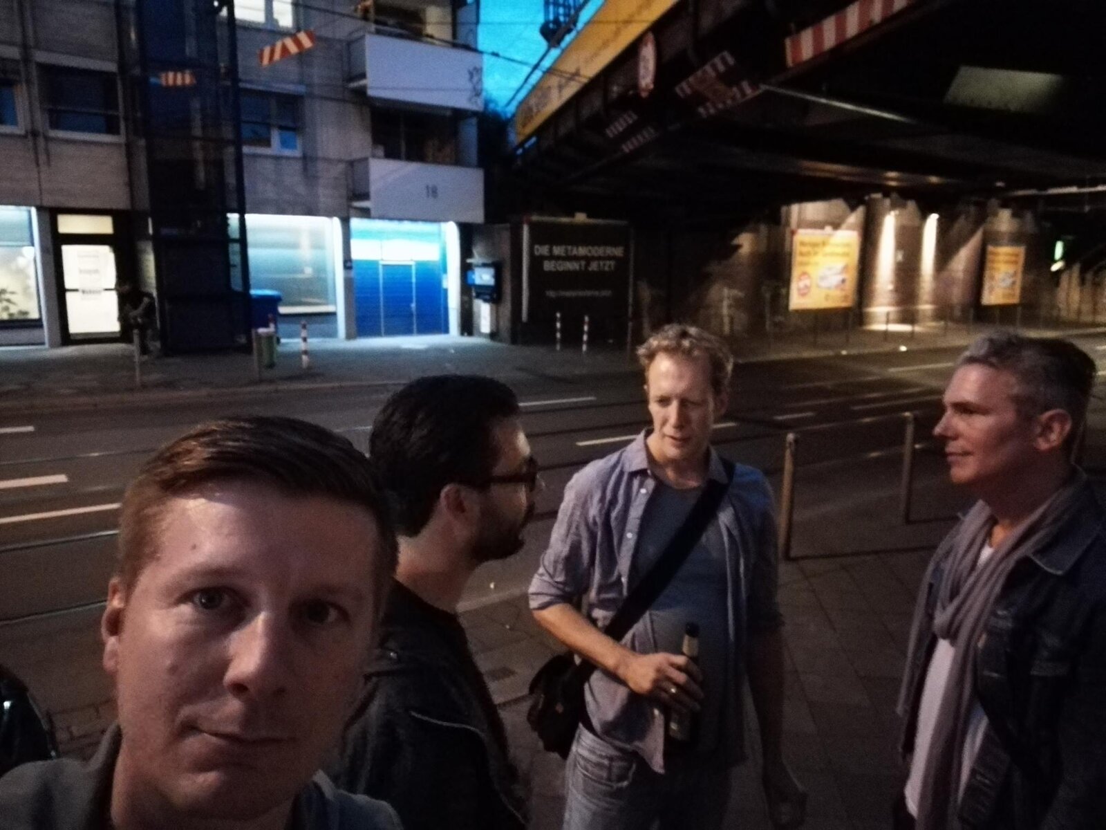 A nighttime street scene near Worringer Platz, four people standing around, the 'DIE METAMODERNE BEGINNT JETZT' poster visible under the railway bridge in the background