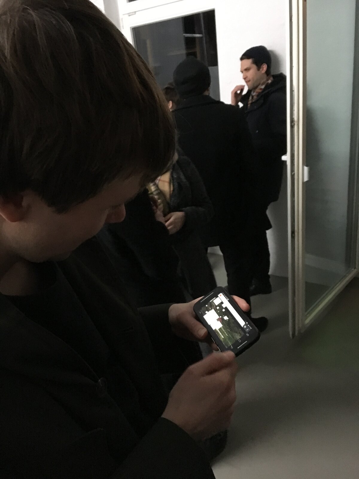 Someone photographing the show on an old smartphone at the Berlin vernissage, a small crowd in the dark hallway behind them