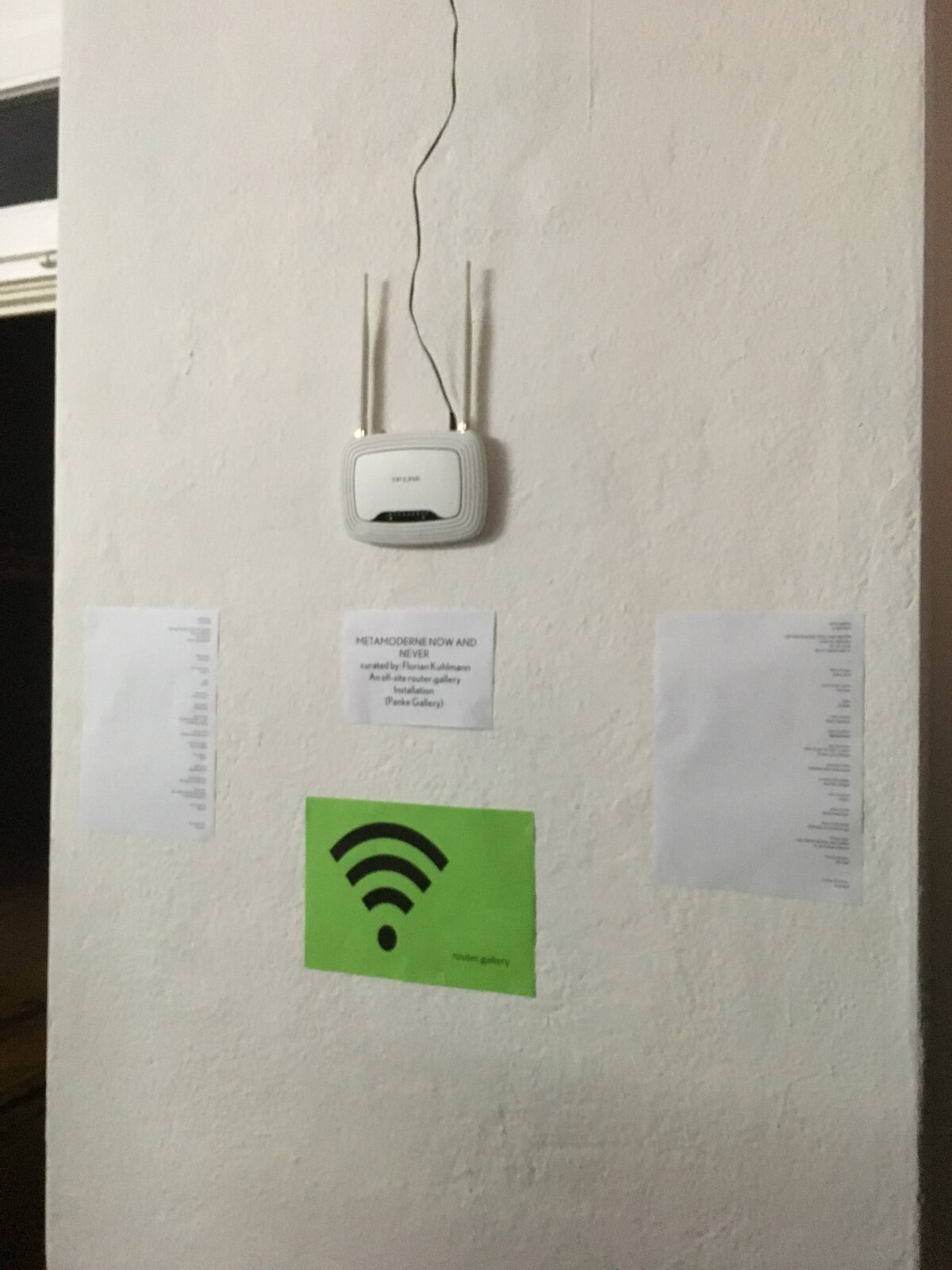 A WLAN router mounted on a gallery wall, with a wall label reading 'METAMODERNE NOW AND NEVER, curated by Florian Kuhlmann, an off-site router.gallery installation, Panke Gallery' and a green wifi symbol below