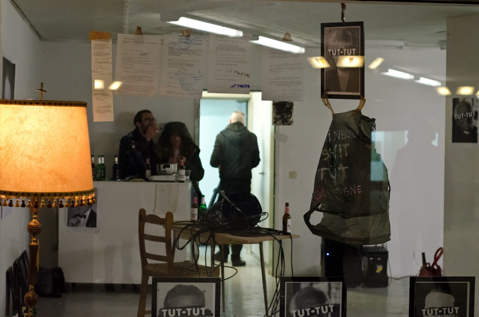 Looking through a glass pane into the shitcologne room: hanging 'TUT-TUT' portraits, a bag reading 'RAINBOW SHIT TUT COLOGNE', handwritten notes taped to the ceiling